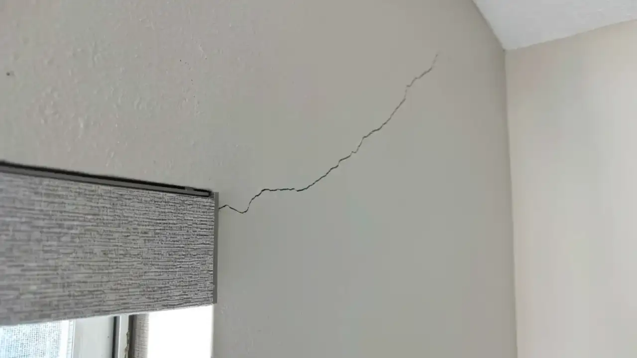 A hairline stress crack running diagonally across a painted interior wall from the head of a window casing up toward the wall-and-ceiling corner