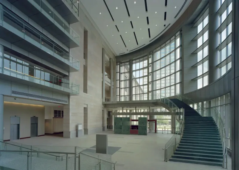 Soaring civic-building atrium in Springfield — a multi-story glass curtain wall, a sweeping stone staircase, and stone-clad walls with smooth plaster-finished surfaces under a finished drywall ceiling