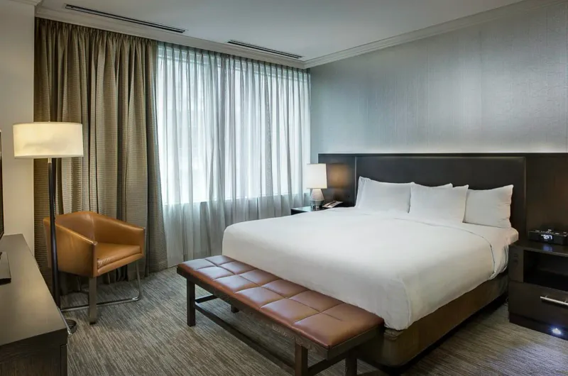 Hotel guest room with finished painted drywall walls and ceiling, a floor lamp, a sheer-curtained window, a leather bench at the foot of the bed, and dark wood nightstands